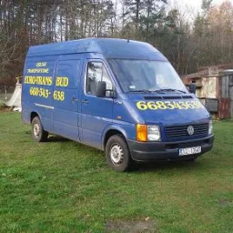 Niebieski van marki Volkswagen z żółtym napisem 'EURO-TRANS BUD' i numerem telefonu 668343638, zaparkowany na trawiastym terenie w pobliżu zardzewiałej szopy